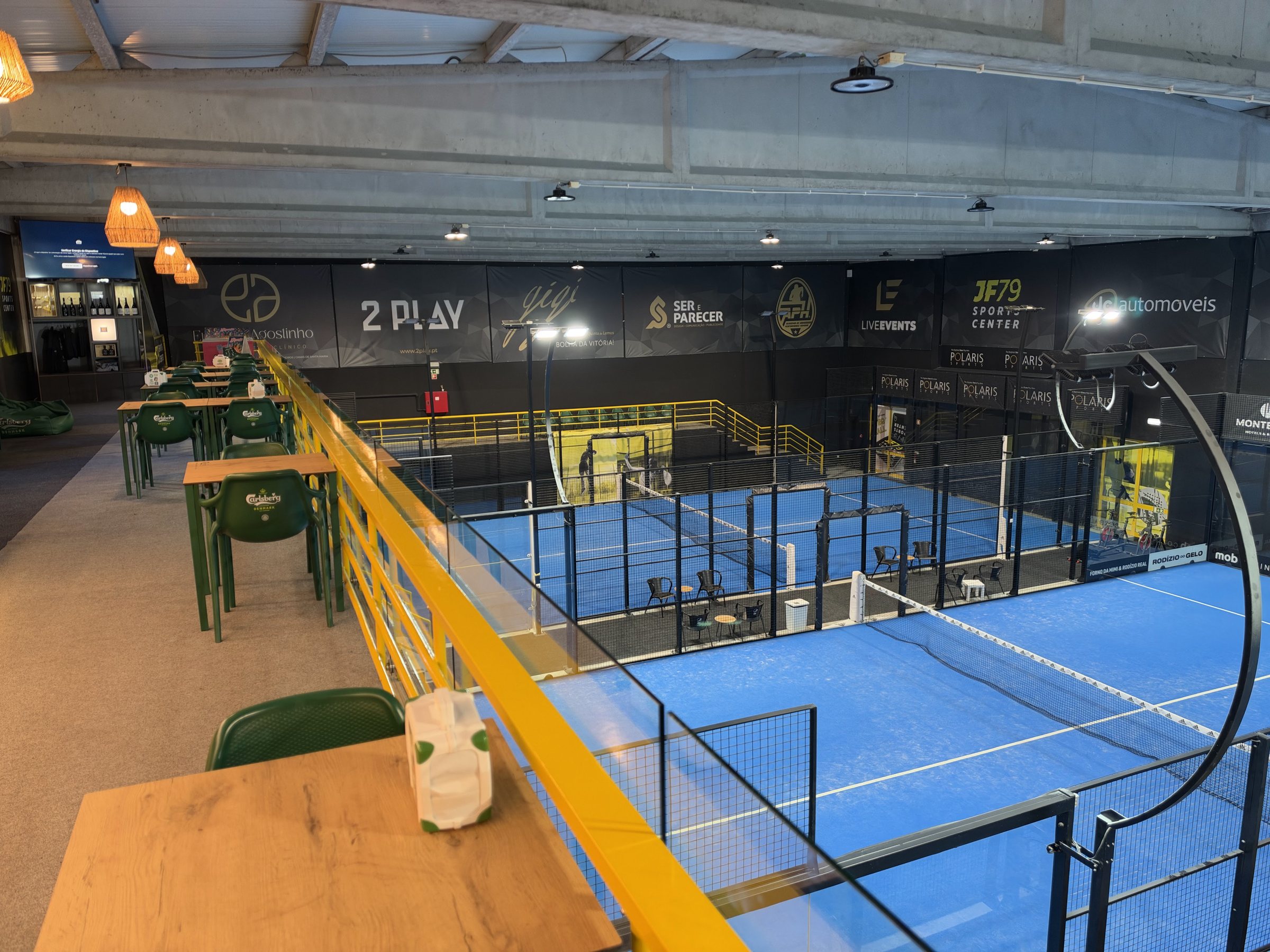 Zona de estar do JF79 Sports Center, com mesas e cadeiras viradas para um campo de padel coberto